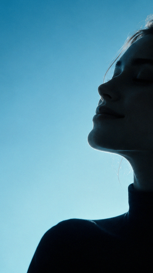 Serene woman with eyes closed, gently tilting her face upward against a blue background.